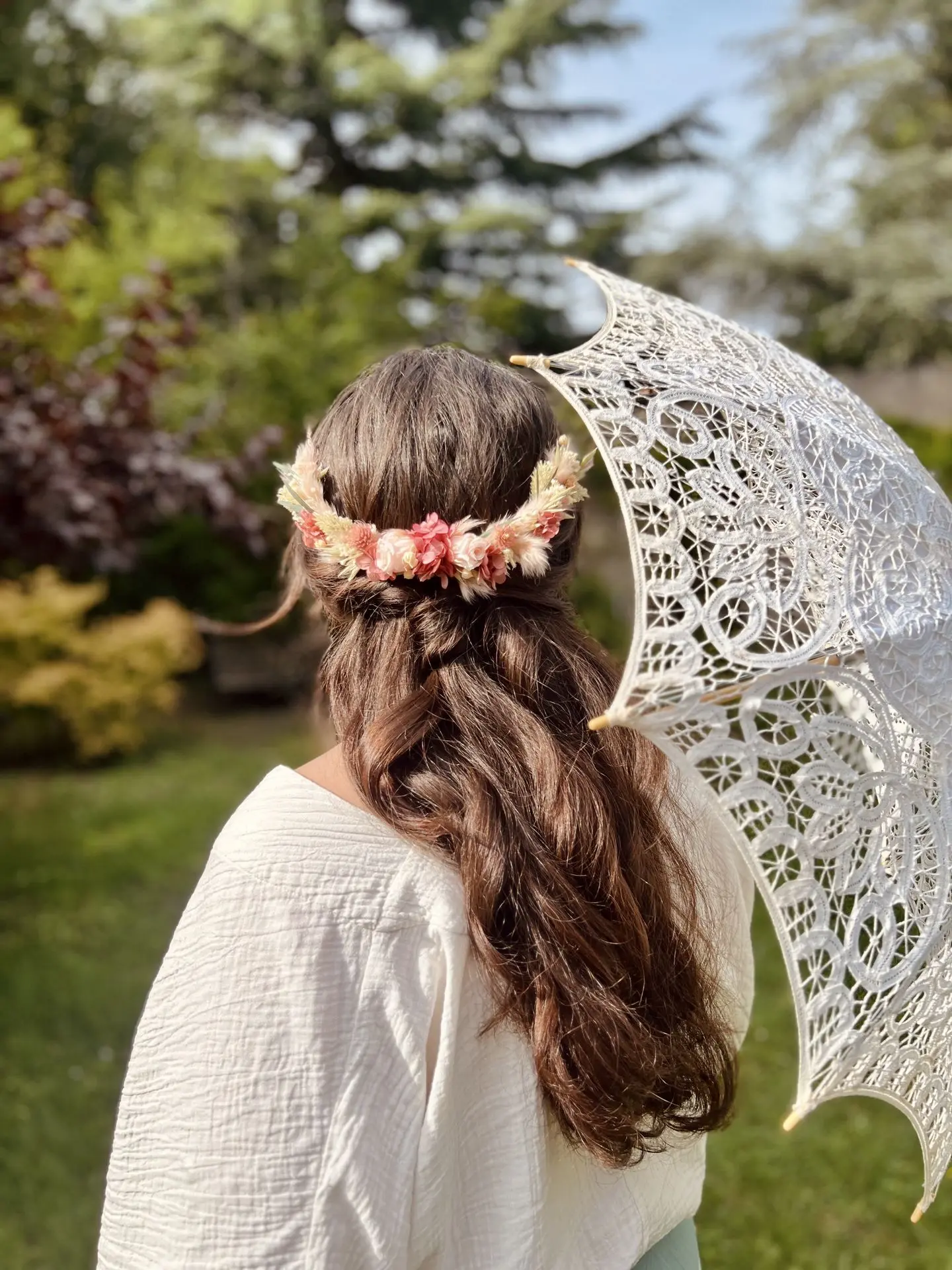 Photo d'une femme de dos portant une couronne de fleurs séchées dans les cheveux et tenant une ombrelle en dentelle